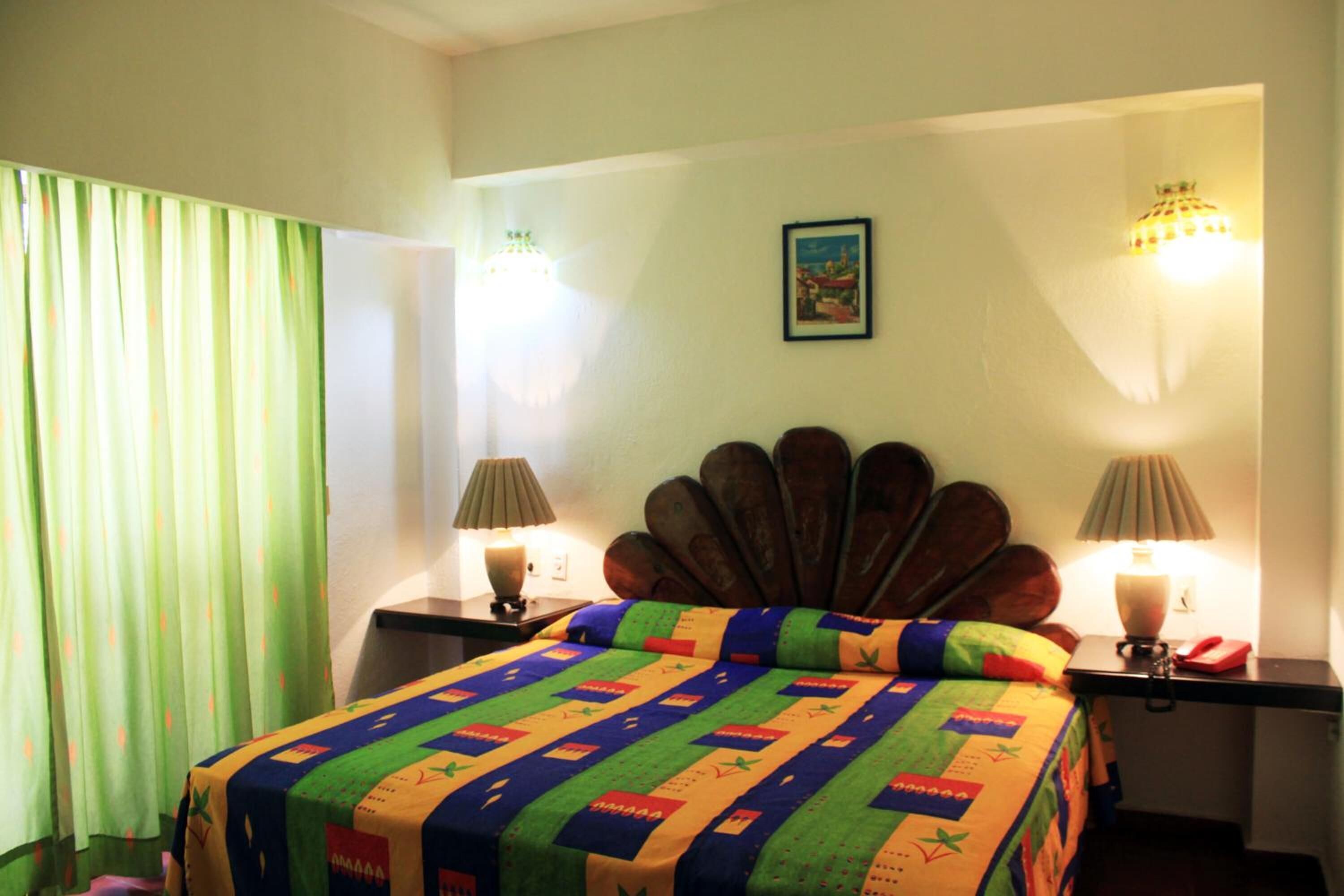 Una camera d'albergo con un letto, due abat-jour, una tenda verde e un quadro appeso al muro.