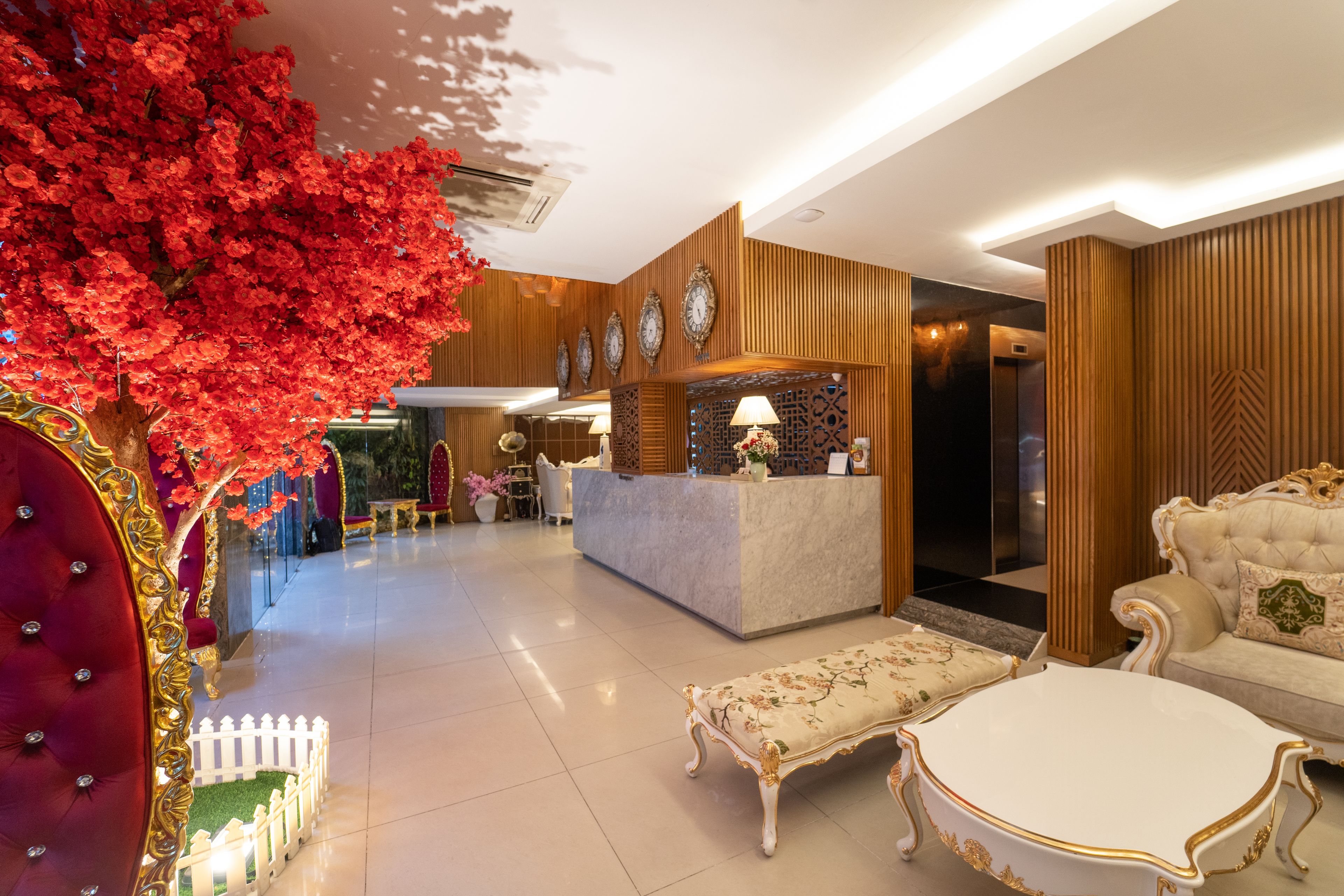 Un vestíbulo de hotel con mostrador de recepción, zona de estar y elementos decorativos como un árbol rojo y una valla blanca.