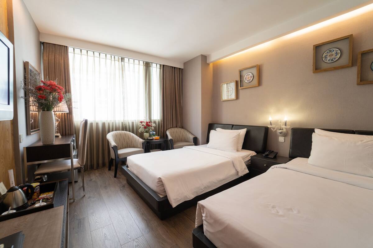 Ein Hotelzimmer mit zwei Betten, einem Schreibtisch, einem Sessel, einem kleinen Tisch mit Blumenvase und einem Fenster mit Vorhängen.