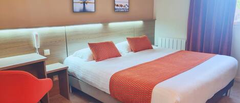 A hotel room with a bed, a desk, a chair, and two framed pictures on the wall.