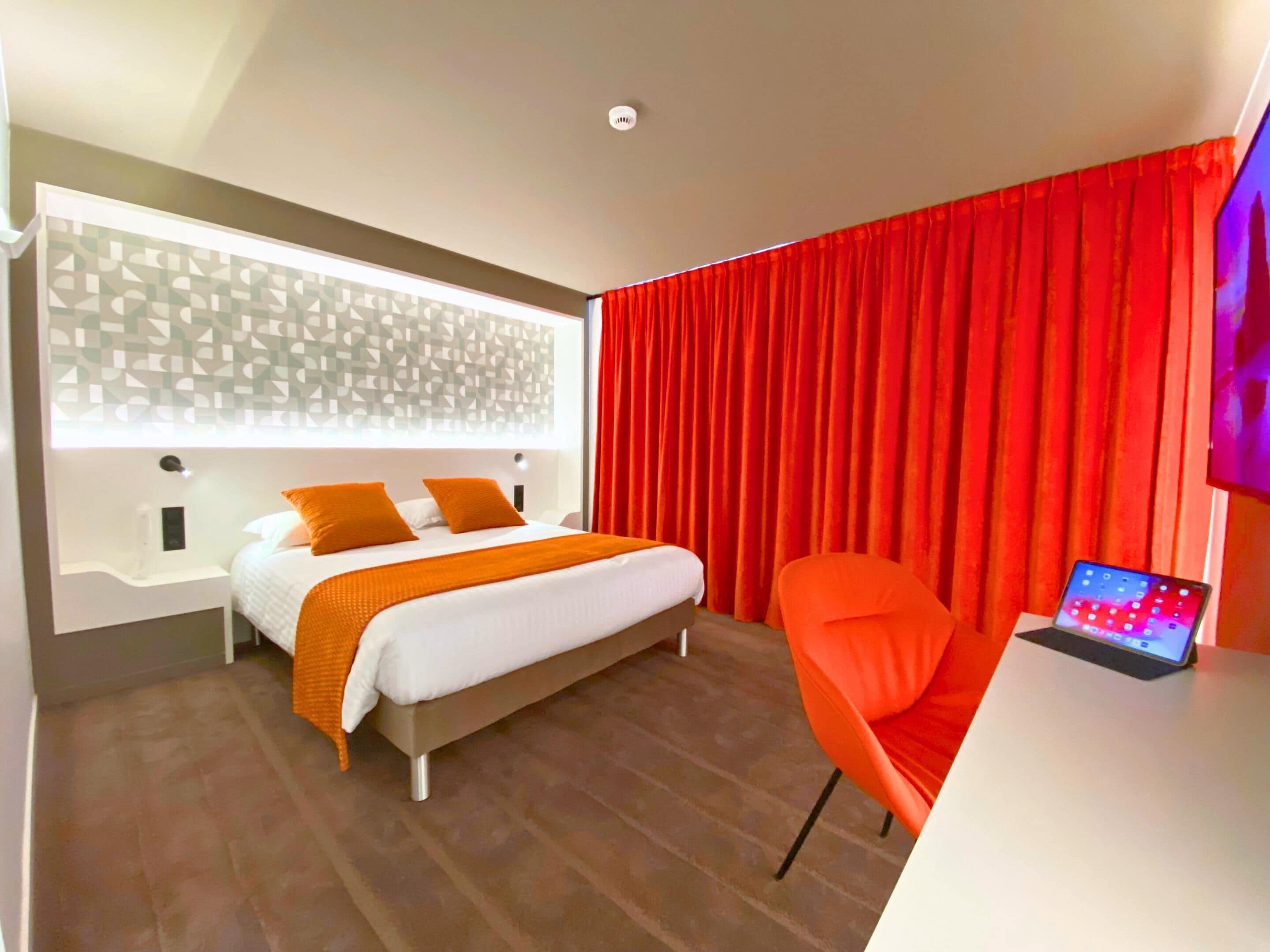 A modern hotel room with a bed, a desk with a laptop, and red curtains.