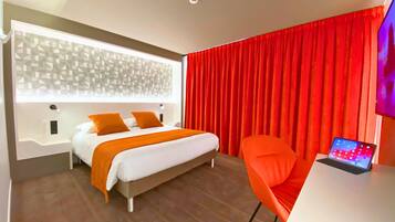 A modern hotel room with a bed, a desk with a laptop, and red curtains.