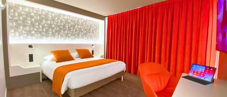 A modern hotel room with a bed, a desk with a laptop, and red curtains.