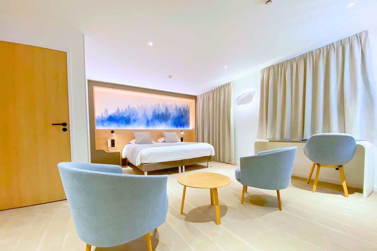 A modern hotel room with a bed, two armchairs, a small table, and a large window with curtains.
