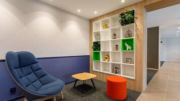 A modern waiting area with a blue cushioned chair, a small wooden table, and a vibrant orange ottoman. A white shelving unit with green and white compartments displays various decorative items.