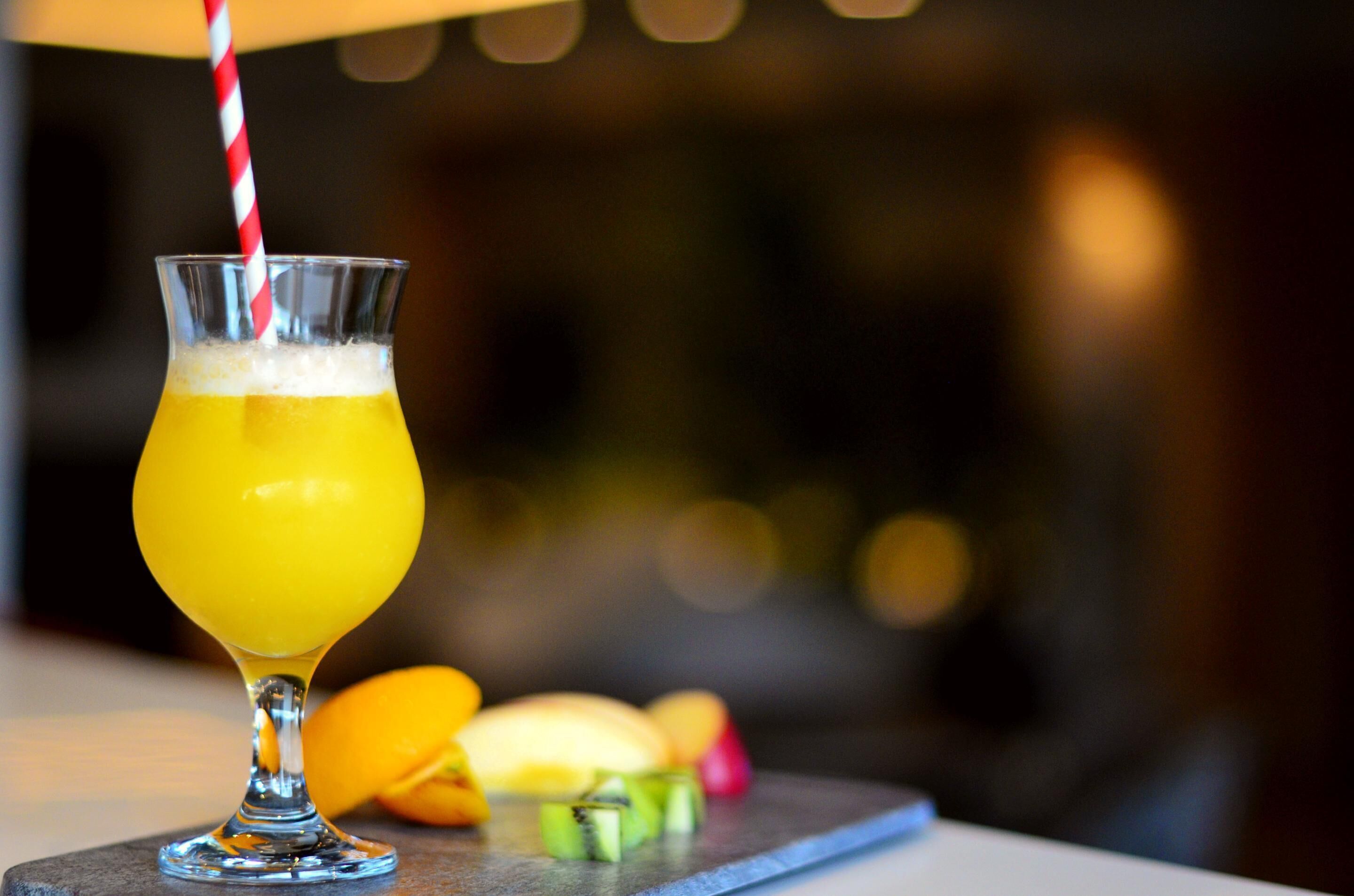 A glass of orange juice with a striped straw, placed on a table with fruits in the background.