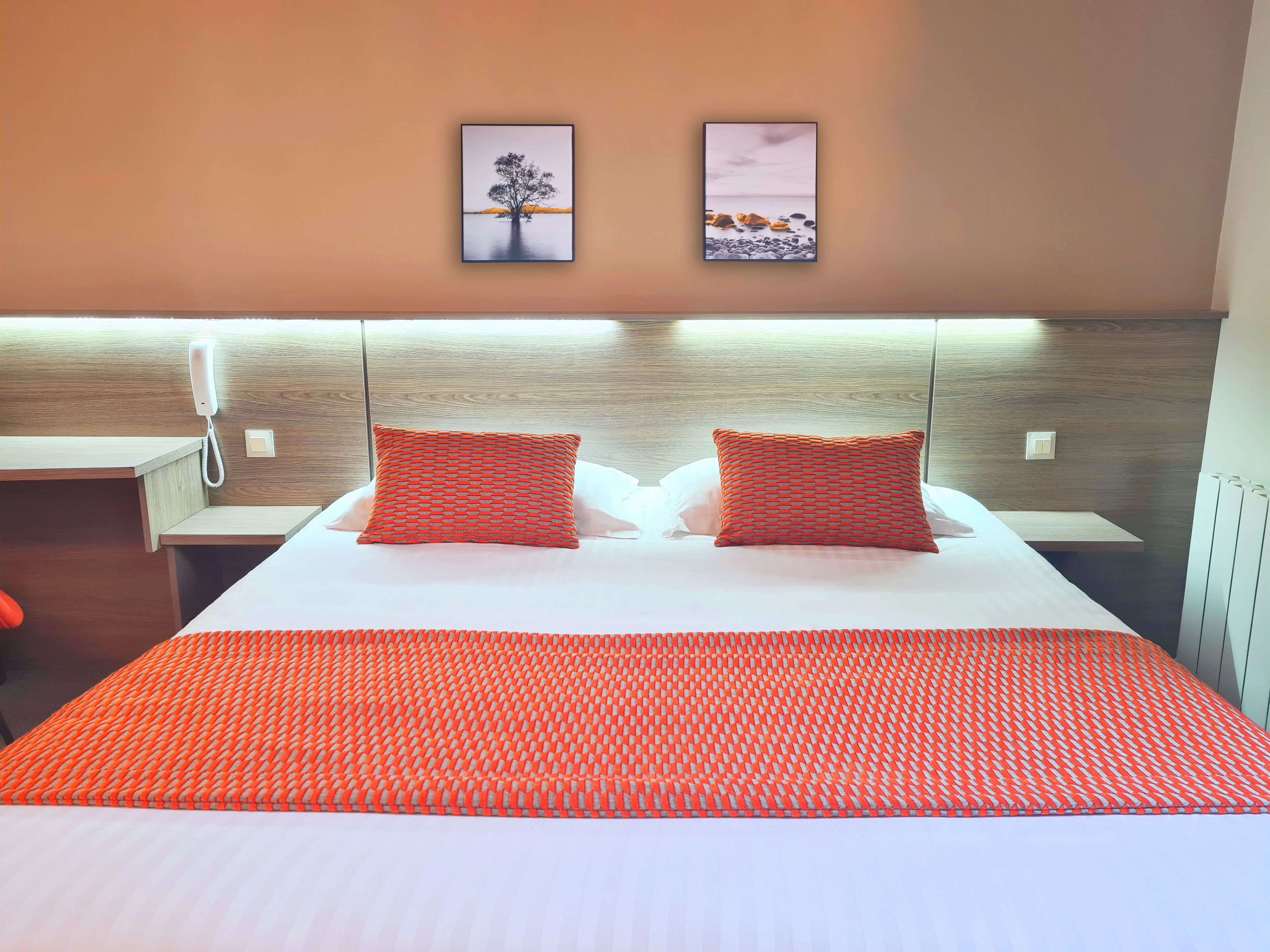 Habitación de hotel con una cama, dos almohadas, una mesita de noche, un teléfono y dos cuadros en la pared.