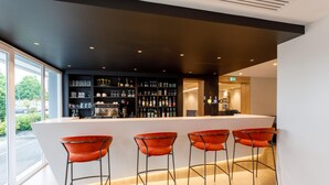 Zona de bar moderna con mostrador blanco, estantes negros repletos de botellas, vasos y una cafetera. Hay cuatro taburetes naranjas.
