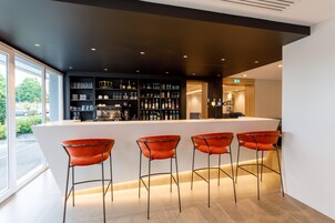 Zona de bar moderna con mostrador blanco, estantes negros repletos de botellas, vasos y una cafetera. Hay cuatro taburetes naranjas.