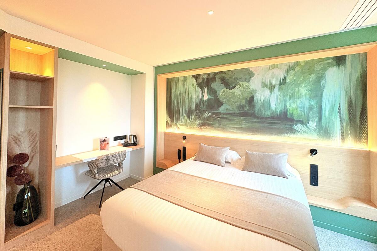 A modern hotel room with a large bed, a desk with a chair, and a painting of a waterfall on the wall.