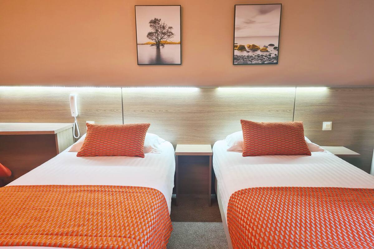 A hotel room with two single beds, each with an orange patterned bedspread and matching pillows. There are two framed pictures on the wall above the beds, one depicting a tree and the other a beach scene.