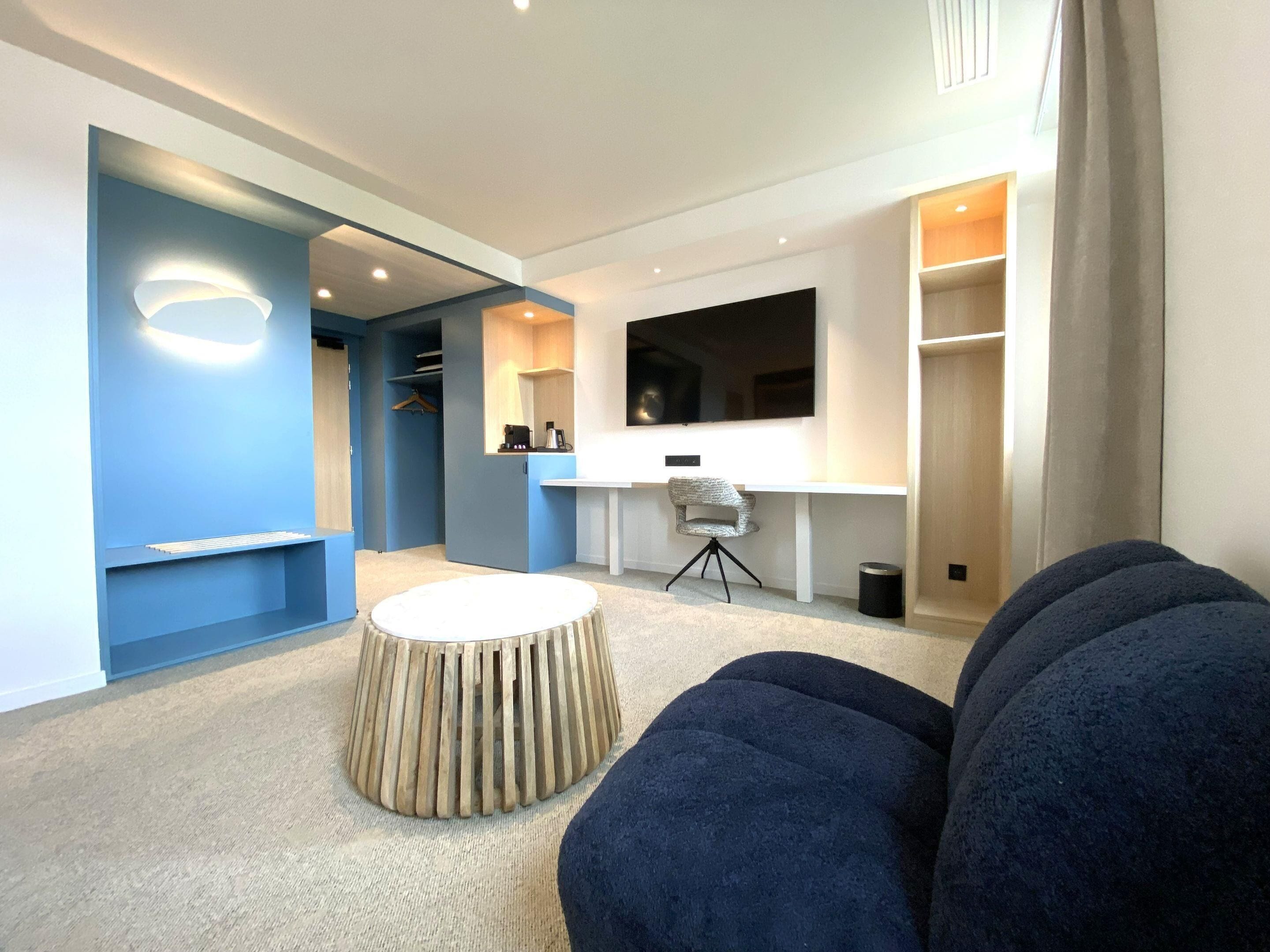 A modern living room with a round wooden coffee table, a dark blue sofa, and a flat-screen TV mounted on the wall.