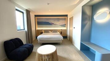 A hotel room with a bed, a round ottoman, a bench, and a wall-mounted light fixture.