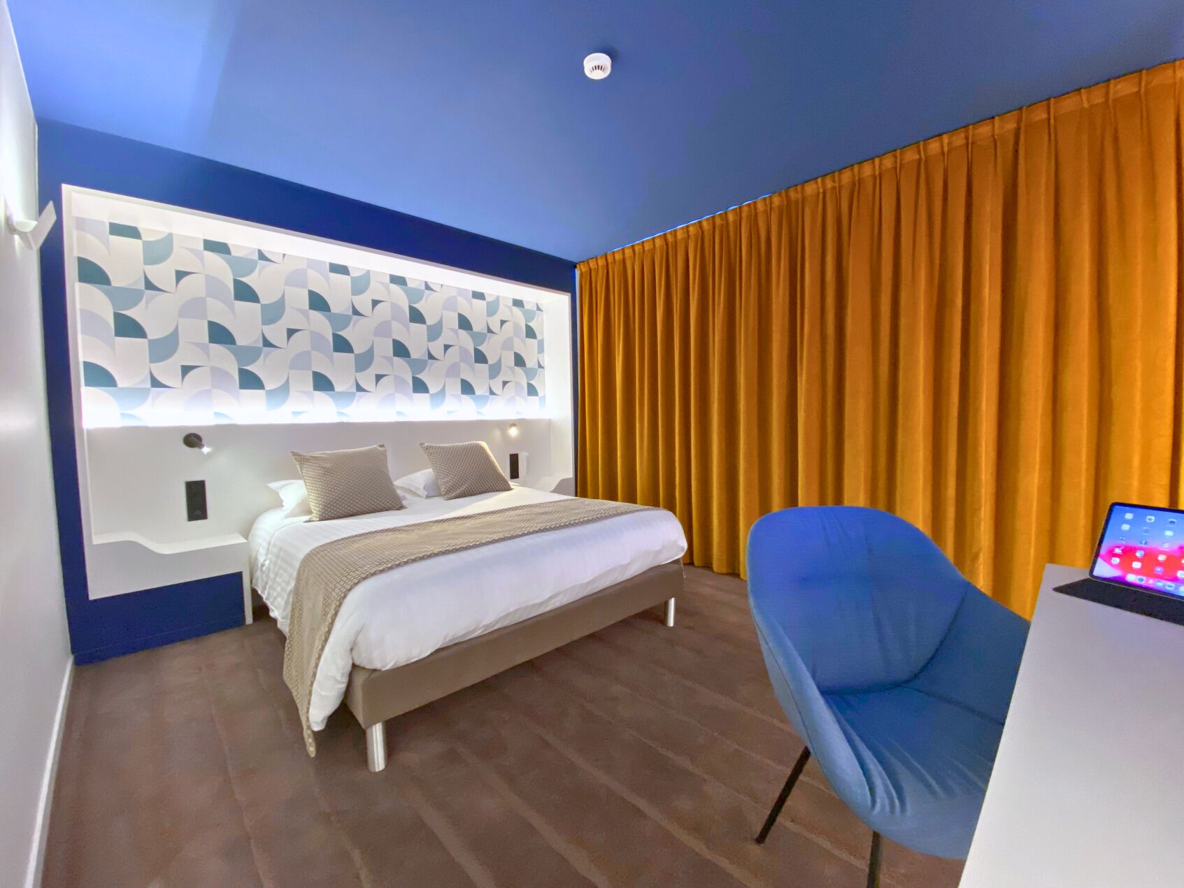 A modern hotel room with a bed, a blue chair, and a laptop on a desk.