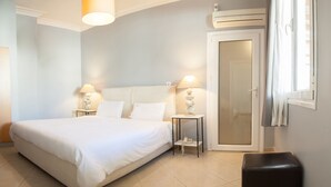 A neatly arranged bedroom with a large bed, bedside lamps, a small table, a door, and a window with curtains. - Kimon Athens Hotel (Athens)