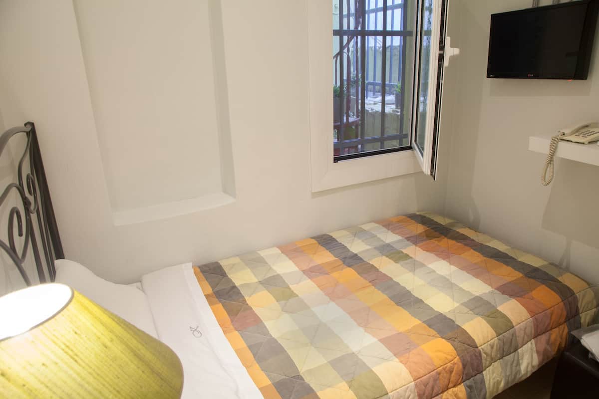 A small, single-bed room with a checkered bedspread, a bedside table, a lamp, a window with bars, and a mounted TV.