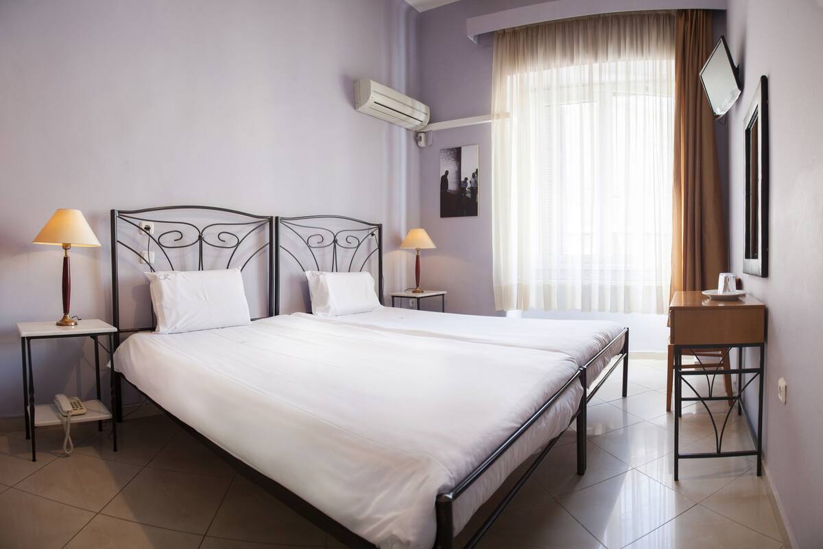 A neatly arranged bedroom with a large bed, two bedside tables with lamps, a small table with a lamp, and a wall-mounted air conditioner.