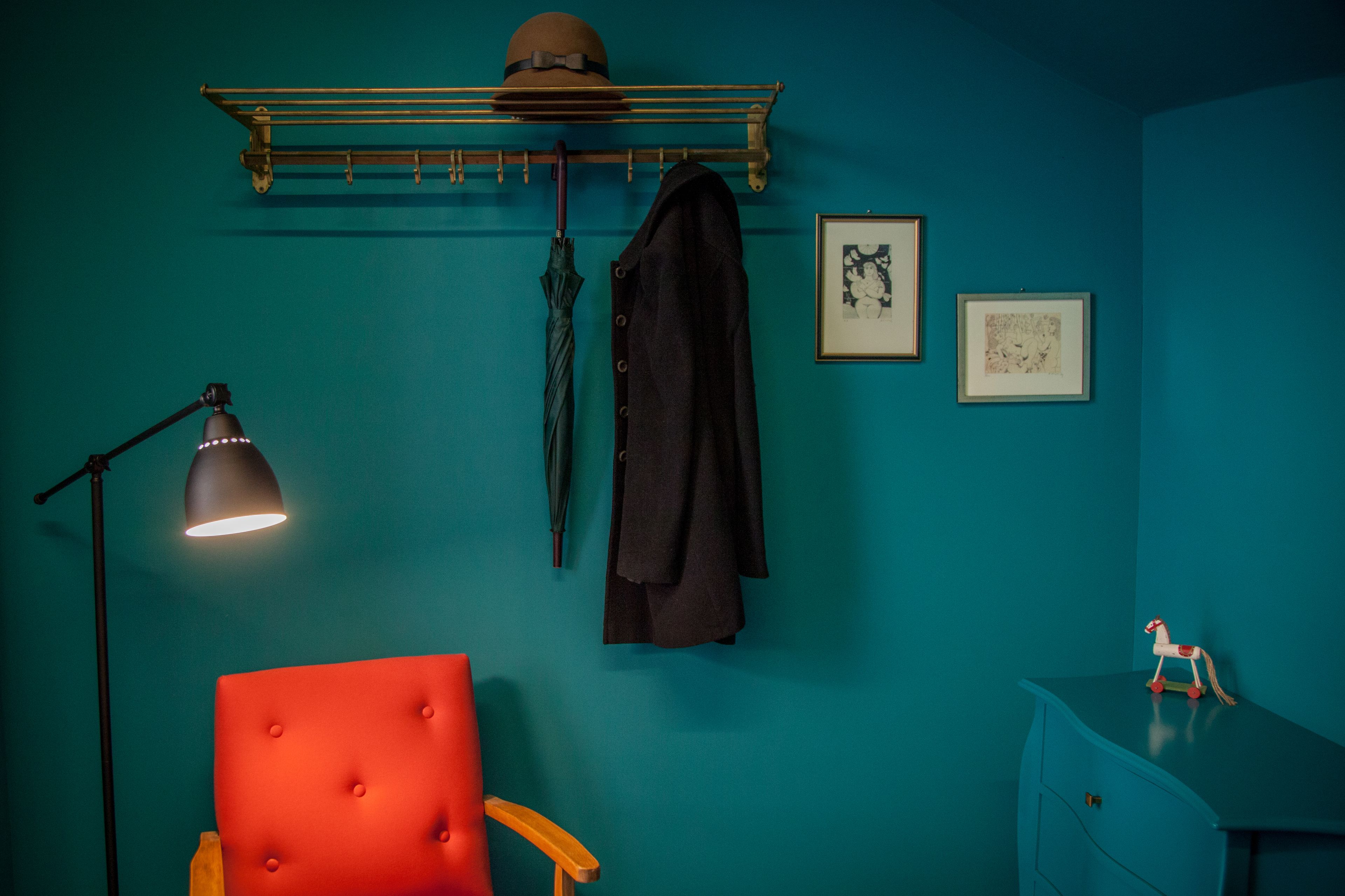 A teal-walled room with a red chair, a black coat, a hat, and a small wooden horse figure on a dresser.