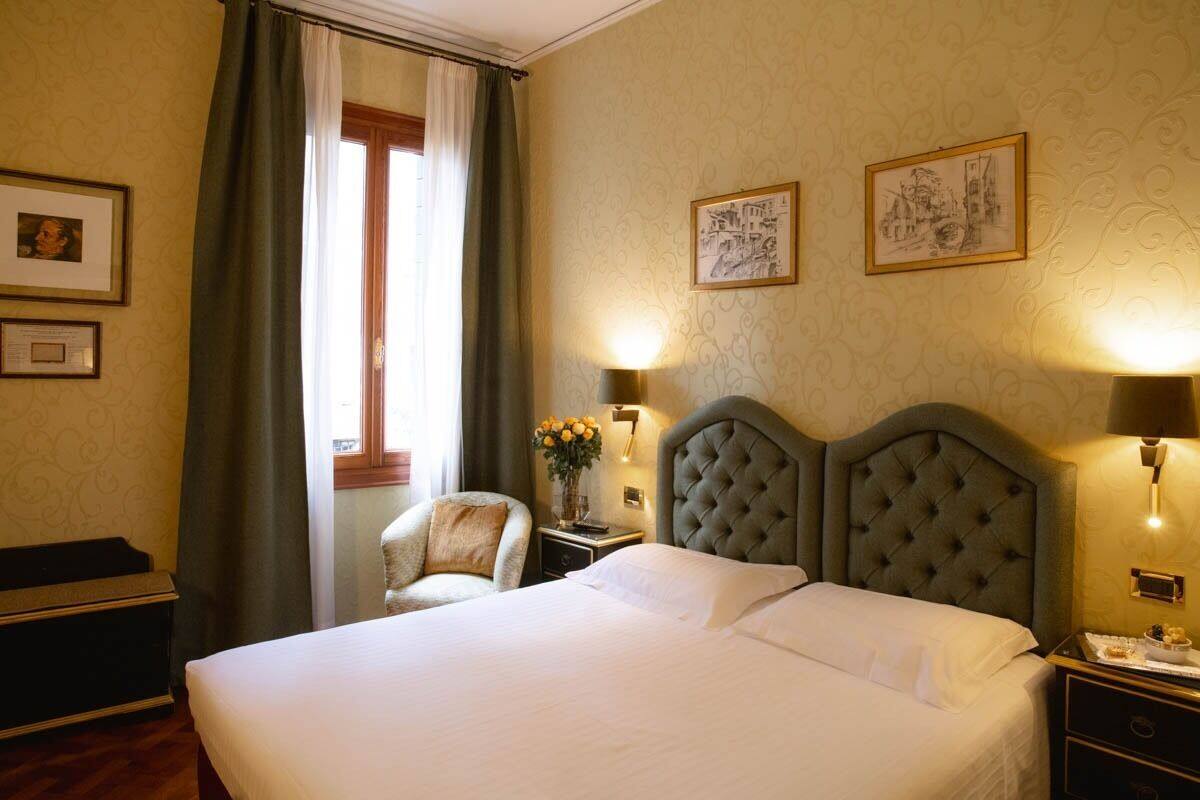 A hotel room with a large bed, two wall-mounted lamps, a chair, and a window with curtains.