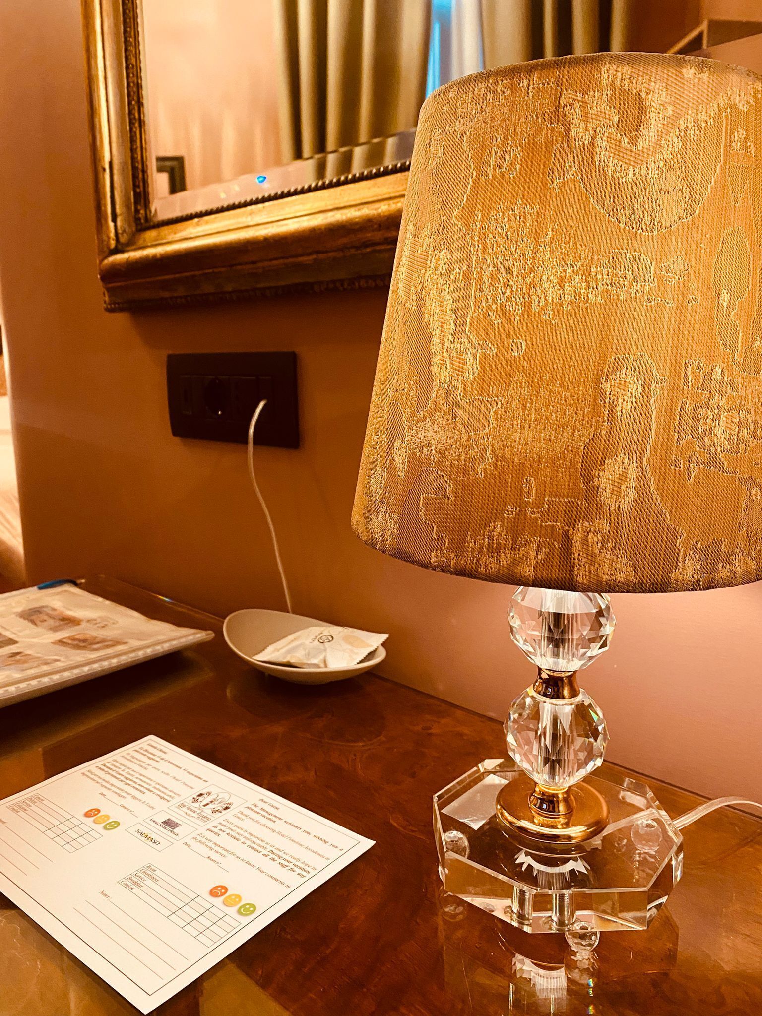 Una habitación de hotel con una mesa de madera, una lámpara con pantalla dorada, un espejo enmarco y un menú sobre la mesa.
