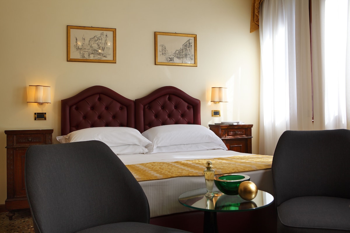 A hotel room with a large bed, two bedside tables, a round glass table with a bowl and a bottle, and two armchairs.