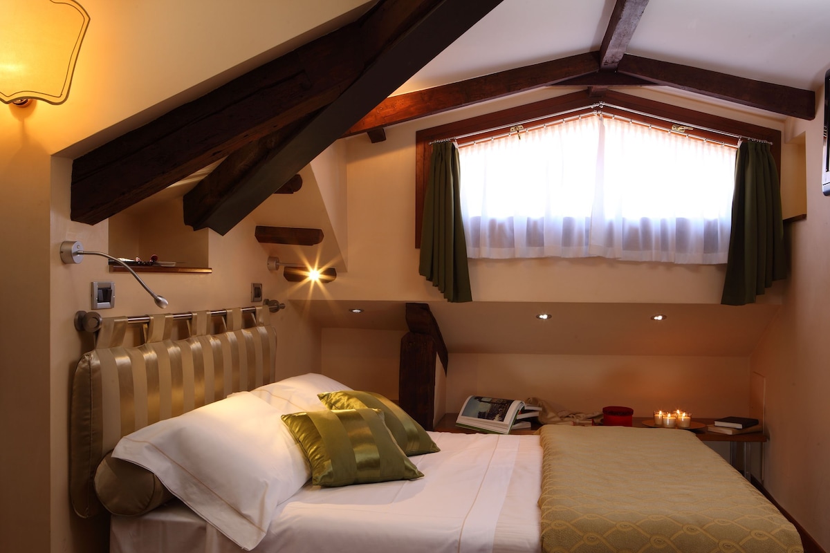 A bedroom with a bed, pillows, a bedside table with a book and candles, a window with curtains, and a radiator.