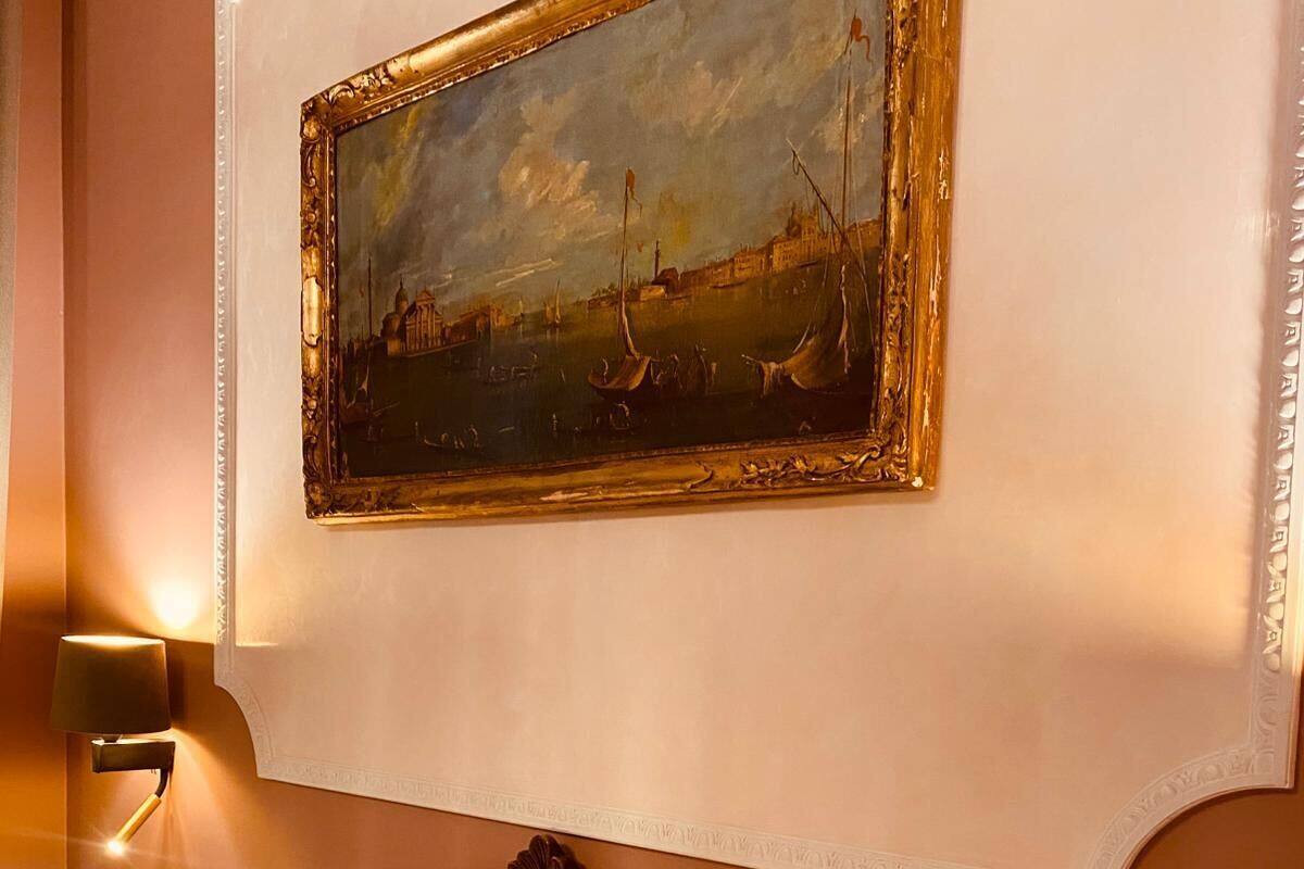 A hotel room with a bed, a painting of a harbor scene, and a wall-mounted light fixture.