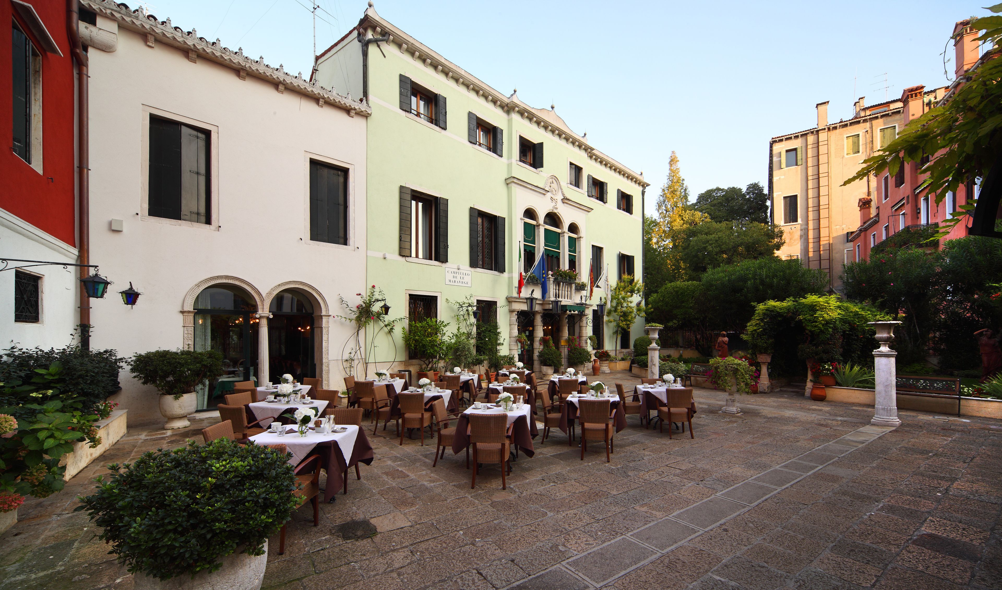 An outdoor café with tables and chairs, surrounded by buildings with balconies and plants.