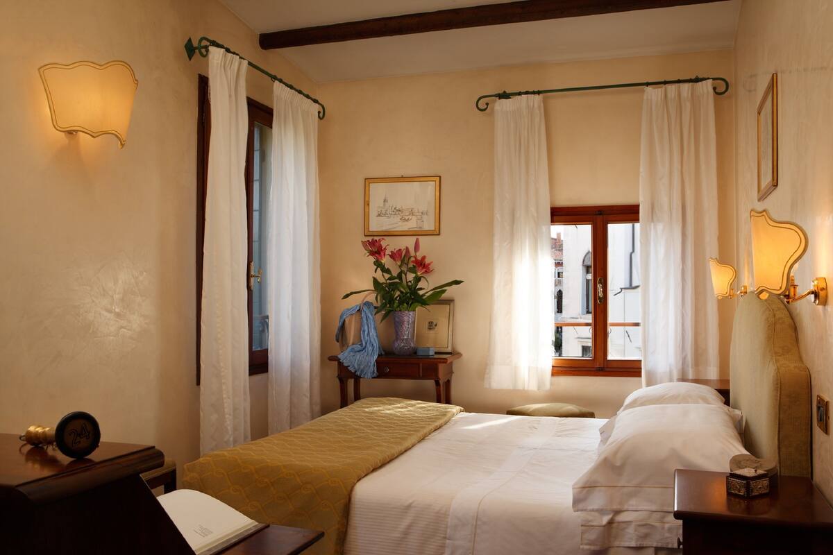 A hotel room with a bed, a nightstand, a desk, a window with curtains, a vase with flowers, and a framed picture on the wall.