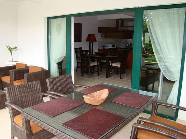A dining area with wicker furniture and a glass table, leading to a kitchen and dining room with wooden chairs and a red lamp. - Hanneman Holiday Residence (Mahé Island)
