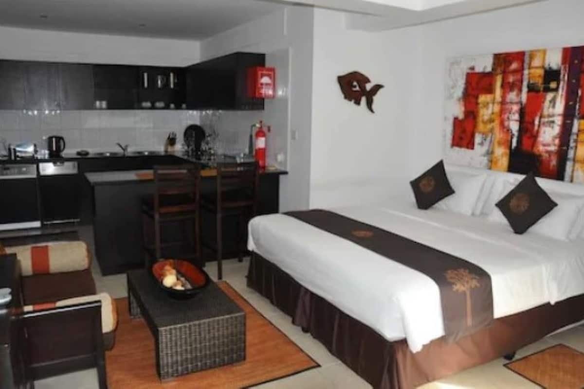 Una habitación de hotel compacta con kitchenette, una cama con cabecera decorativa y un pequeño rincón de descanso.