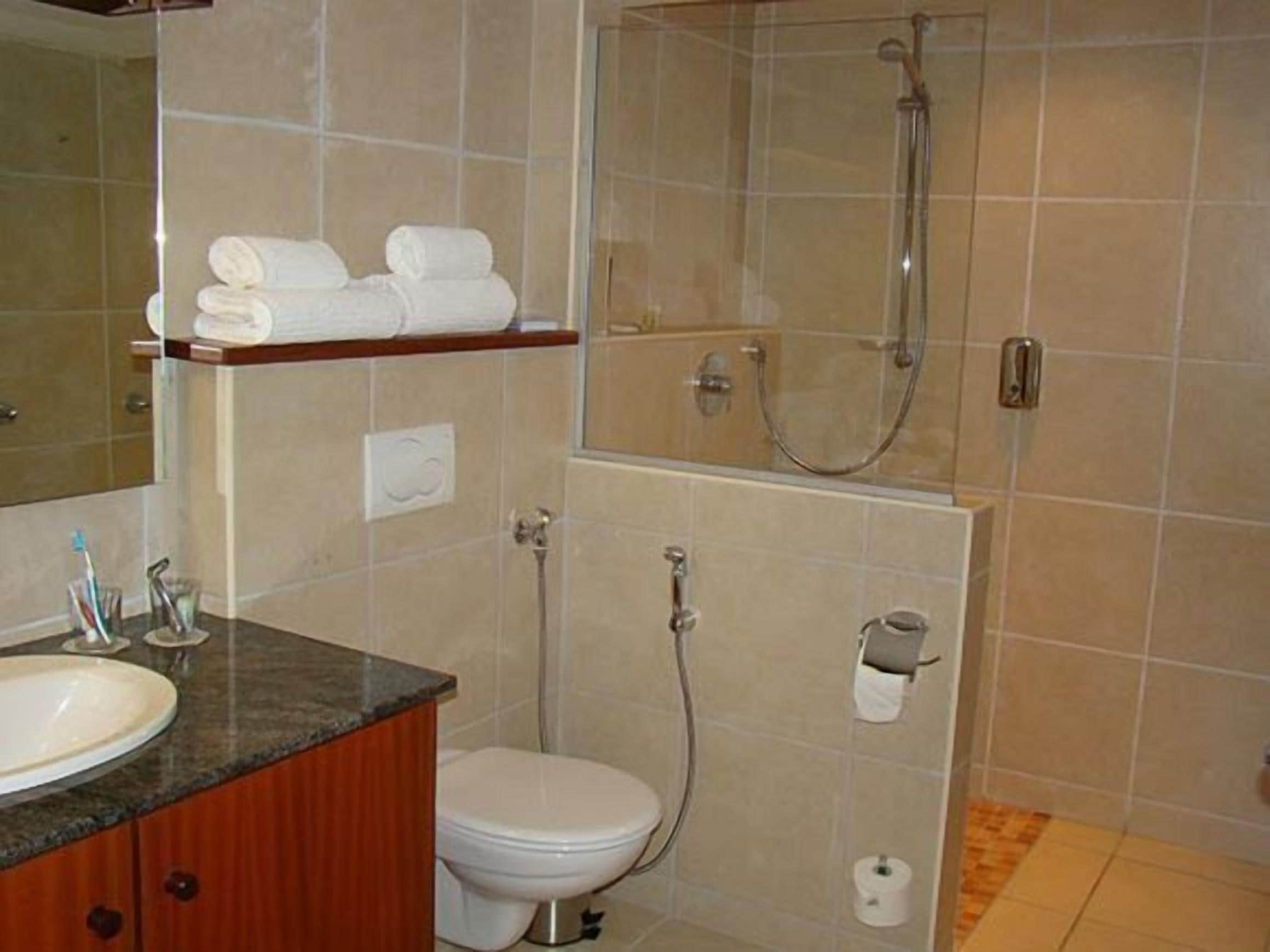 Ein Badezimmer mit Waschbecken, Toilette und Dusche. Handtücher liegen auf einem Regal und eine Toilettenpapierhalterung ist vorhanden.