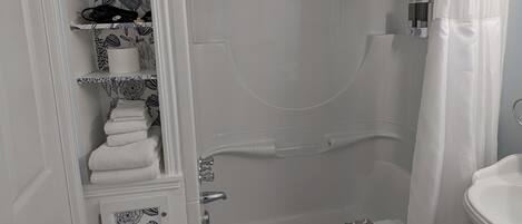 Un baño con bañera, ducha y lavamanos. Hay repisas con toallas y artículos de aseo.