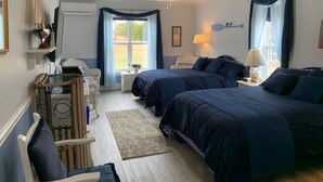 Une chambre avec deux lits, un ventilateur de plafond, une fenêtre avec des rideaux, une petite table avec une lampe et une chaise en osier.