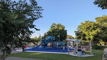 Une aire de jeux avec des toboggans et des structures d’escalade, entourée d’arbres et comprenant un espace pique-nique.