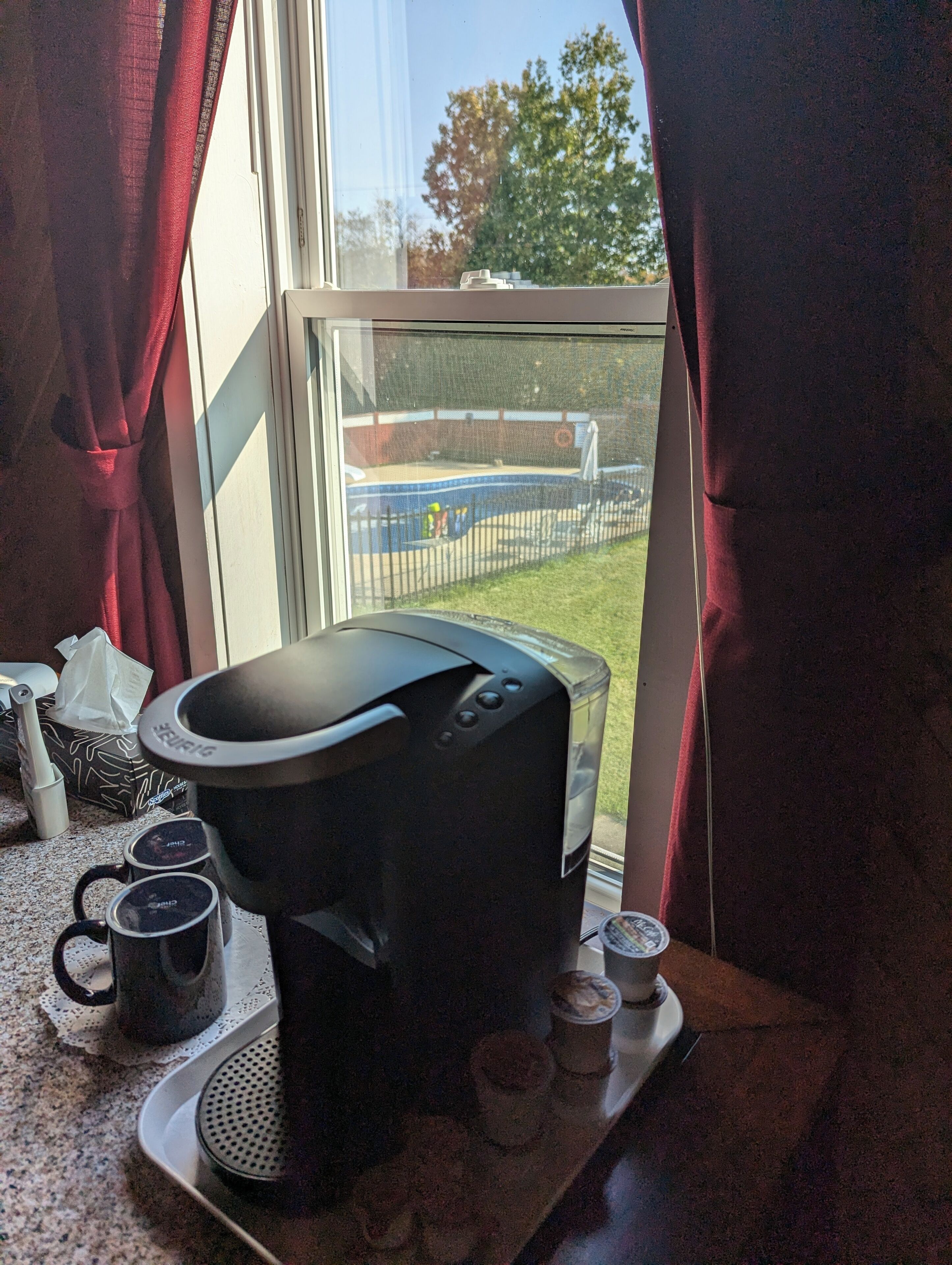 Una cafetera sobre una encimera de cocina con vistas a una piscina y una valla exterior.
