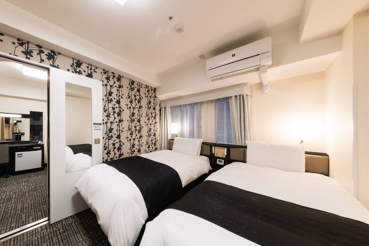 A hotel room with two beds, a mirror, a desk, a wardrobe, an air conditioning unit, and a window with curtains.
