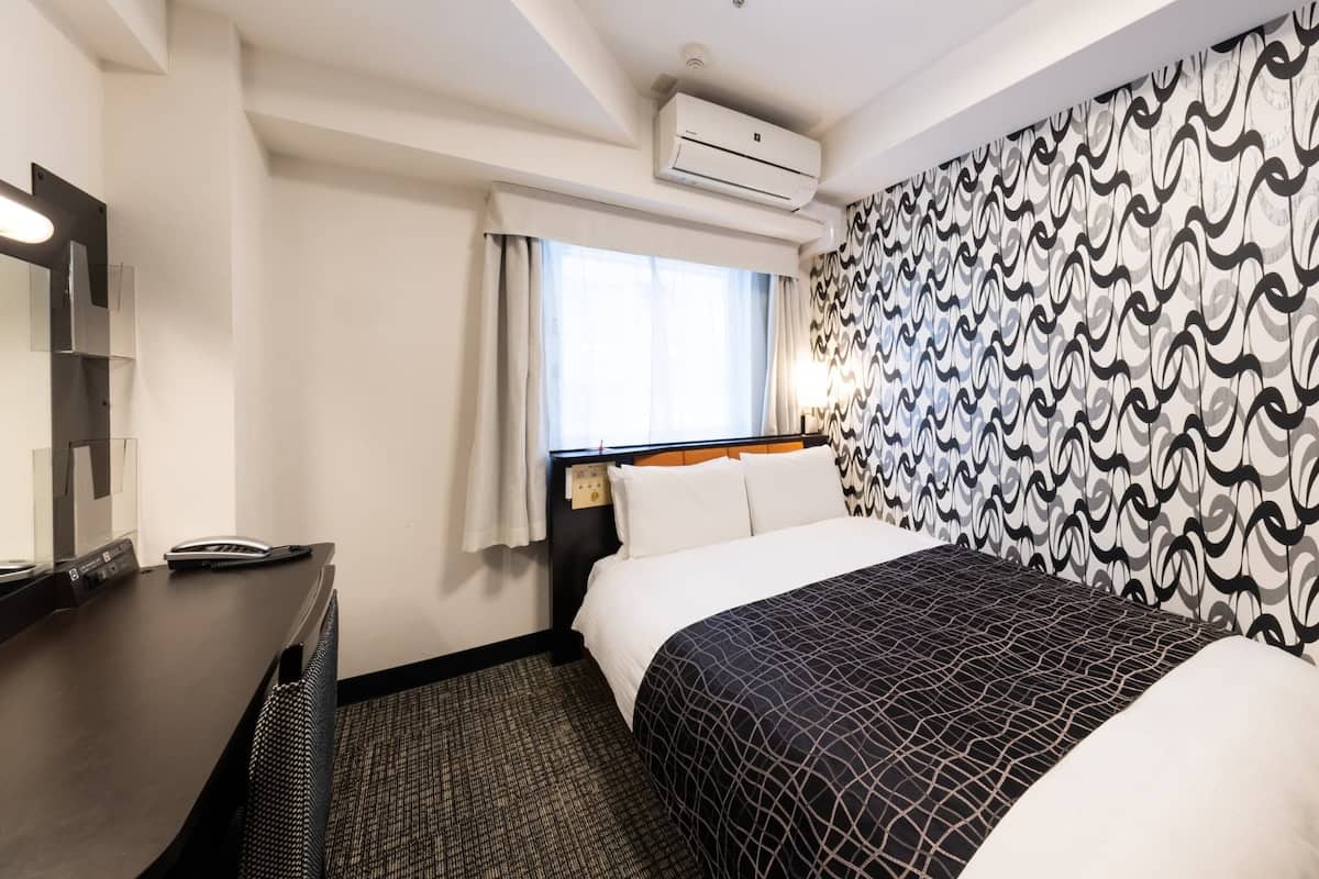 A hotel room with a bed, a desk, a mirror, an air conditioning unit, and a patterned wall.