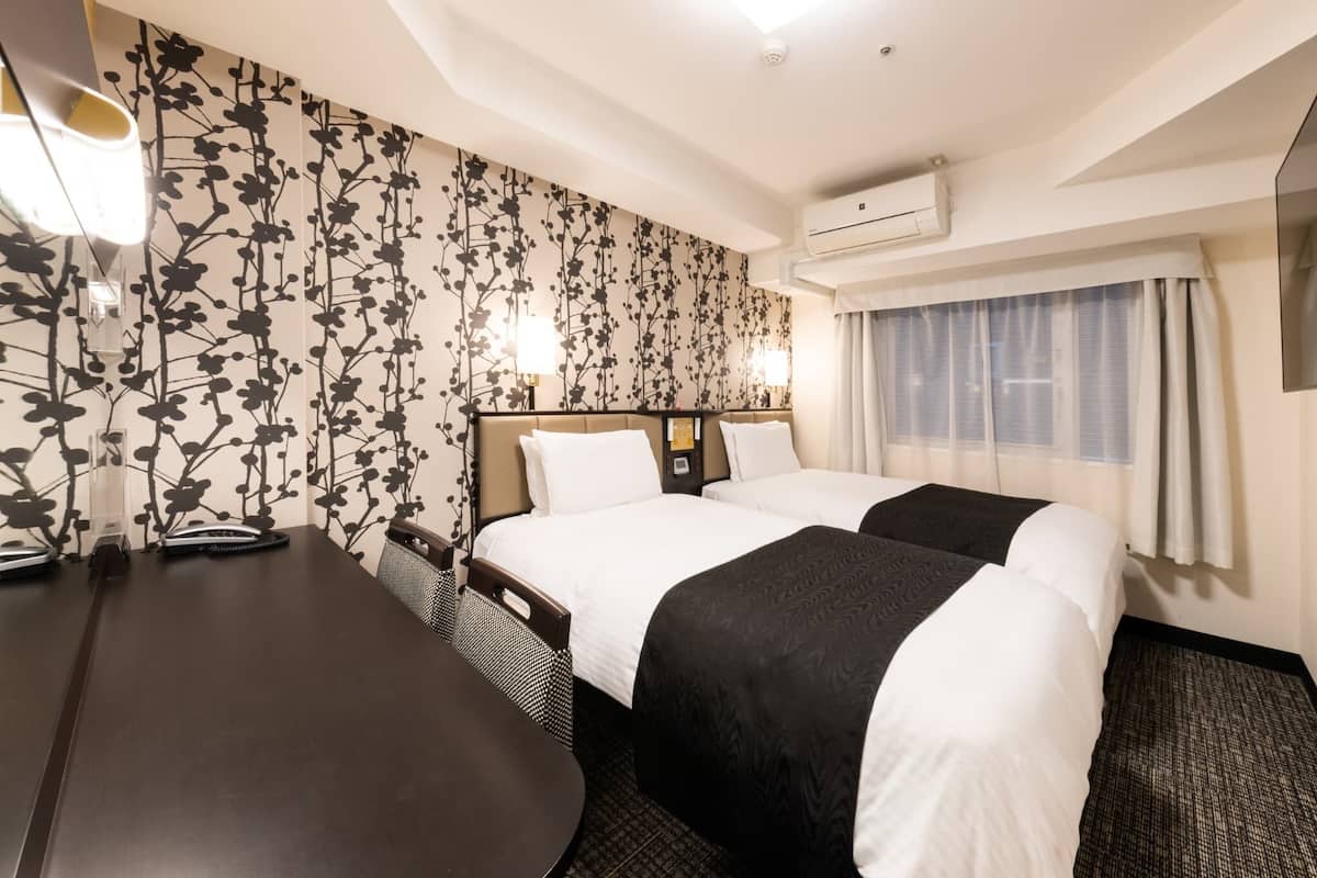 A hotel room with two beds, a desk with a chair, a large window with curtains, and a floral wall design.