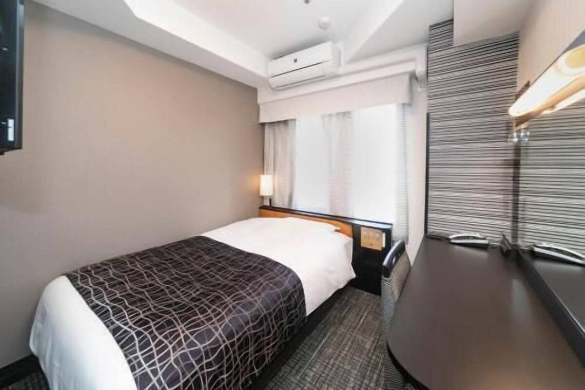 A hotel room with a bed, bedside table, lamp, and a view of the outside.