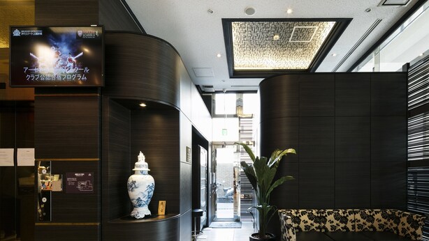 Un lobby moderno con un mostrador de recepción curvo, un gran jarrón decorativo, un sofá con tapicería floral y una pantalla de televisión mostrando un anuncio.