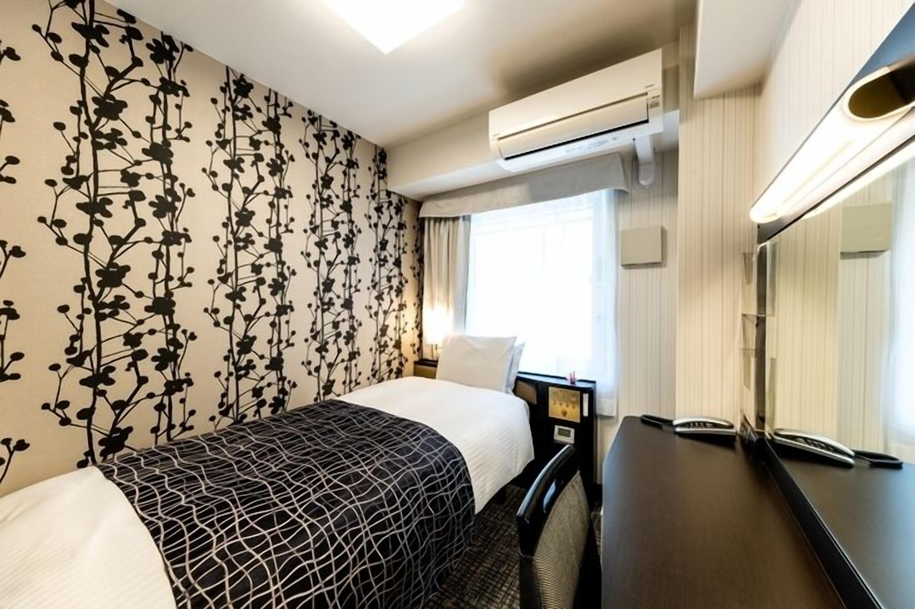 A hotel room with a bed, a desk with a telephone, an air conditioning unit, and a window with curtains.