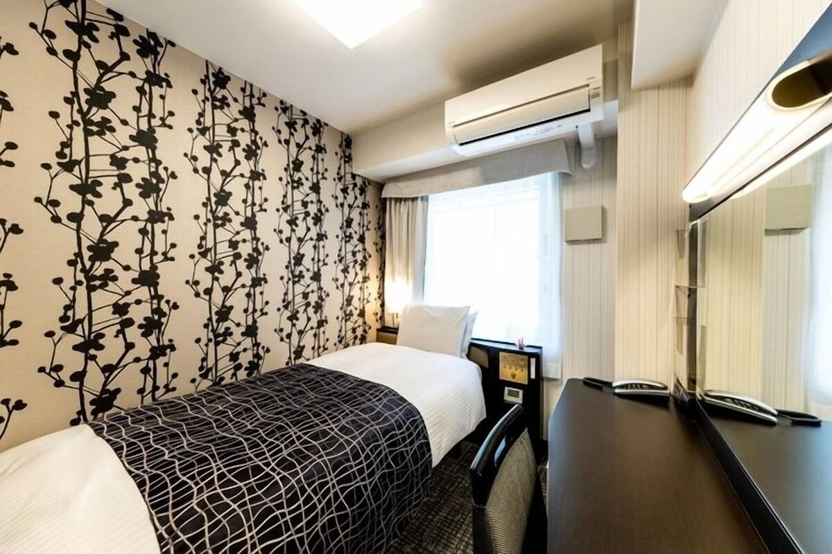 Habitación de hotel con cama, escritorio, teléfono, aire acondicionado y ventana con cortinas.