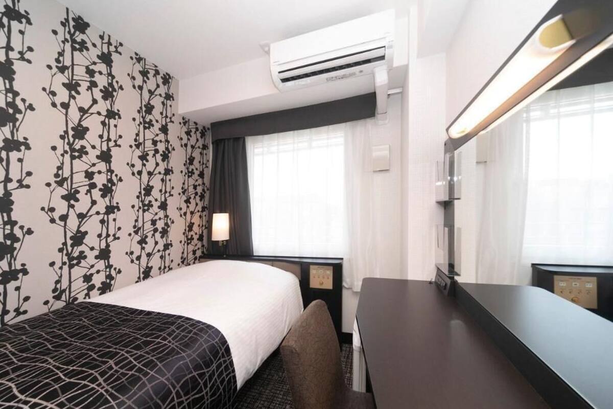 Habitación de hotel con cama, escritorio, silla, espejo, equipo de aire acondicionado y ventana con cortinas.