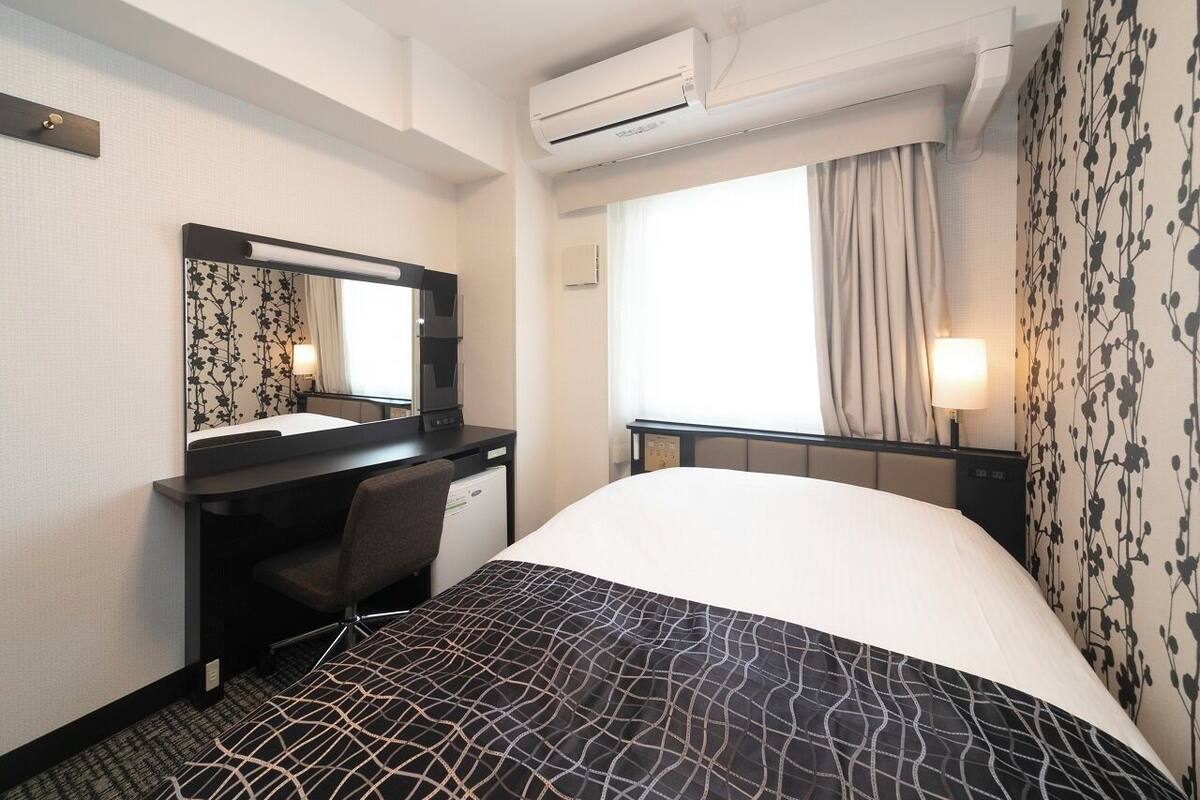 Una habitación de hotel con cama, escritorio con silla, aire acondicionado, ventana con cortinas y pared con estampado floral.