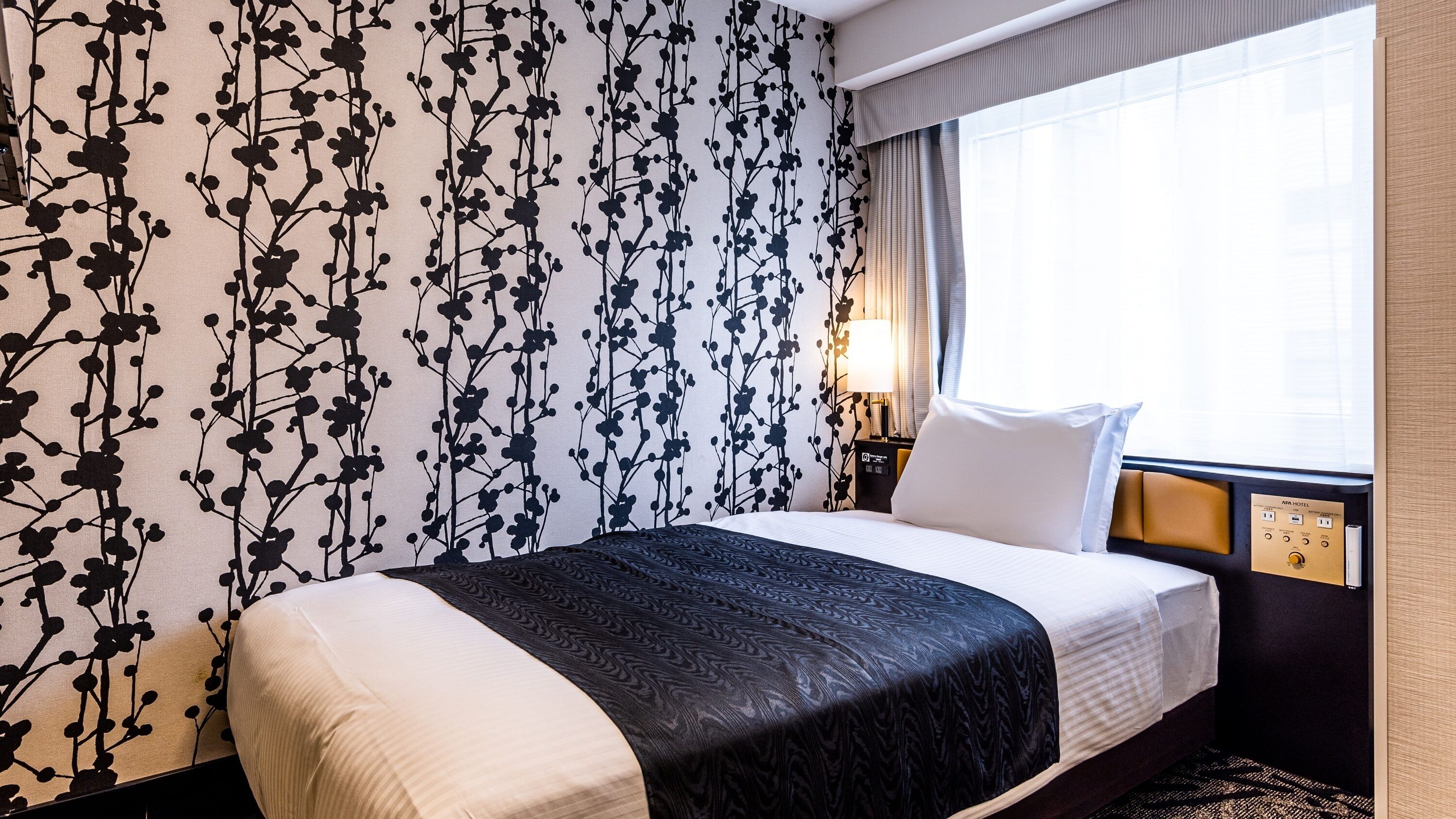 A hotel room with a bed, a nightstand, a lamp, and a window with curtains.