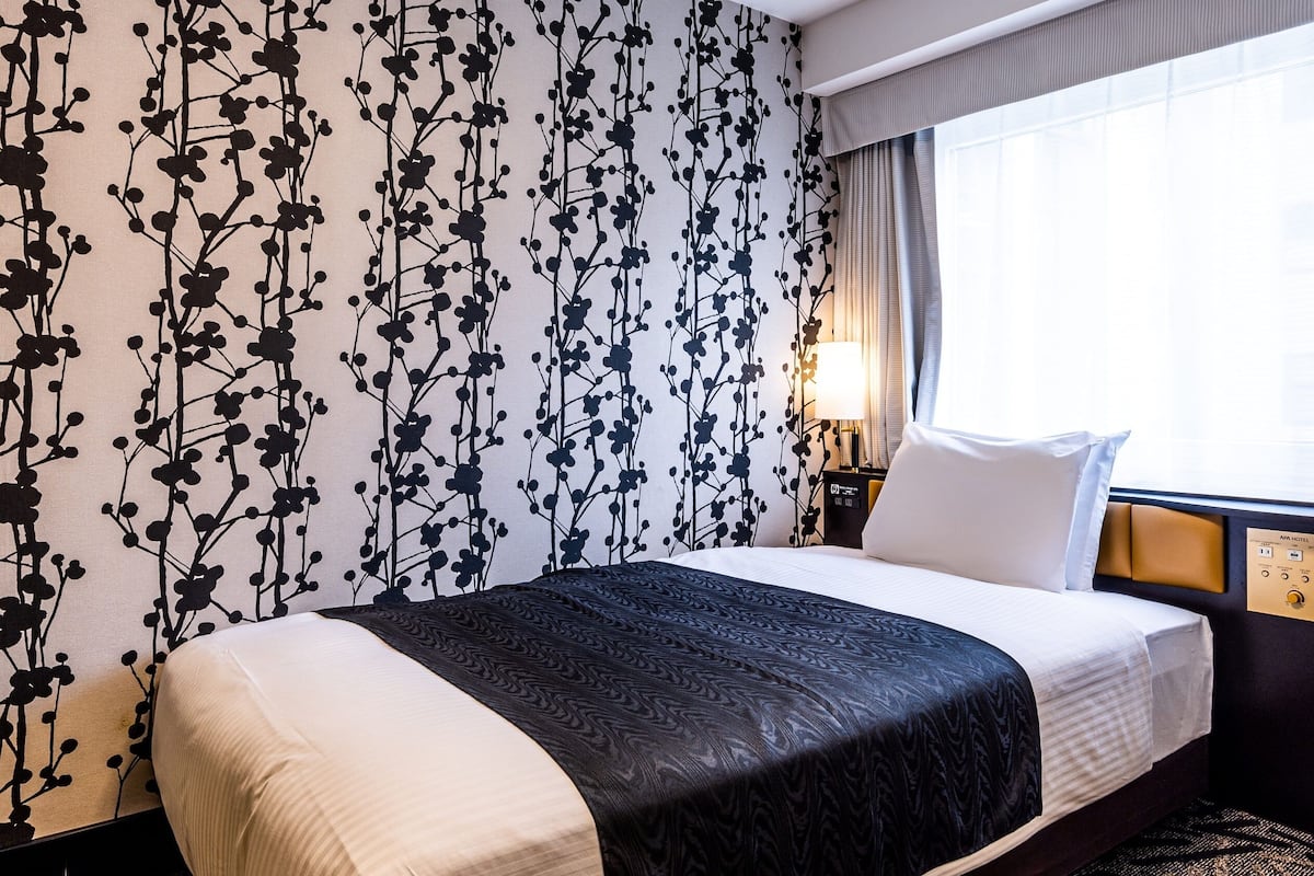 A hotel room with a bed, a nightstand, a lamp, and a window with curtains.