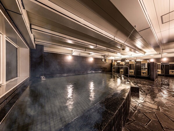 Une piscine intérieure moderne, dotée d’un sol carrelé en pierre, entourée de murs sombres et équipée de plusieurs douches.