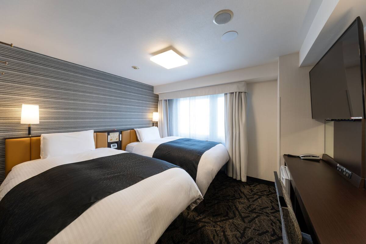 Ein Hotelzimmer mit zwei Betten, einem Flachbildfernseher, einem Schreibtisch und einem Fenster mit Vorhängen.