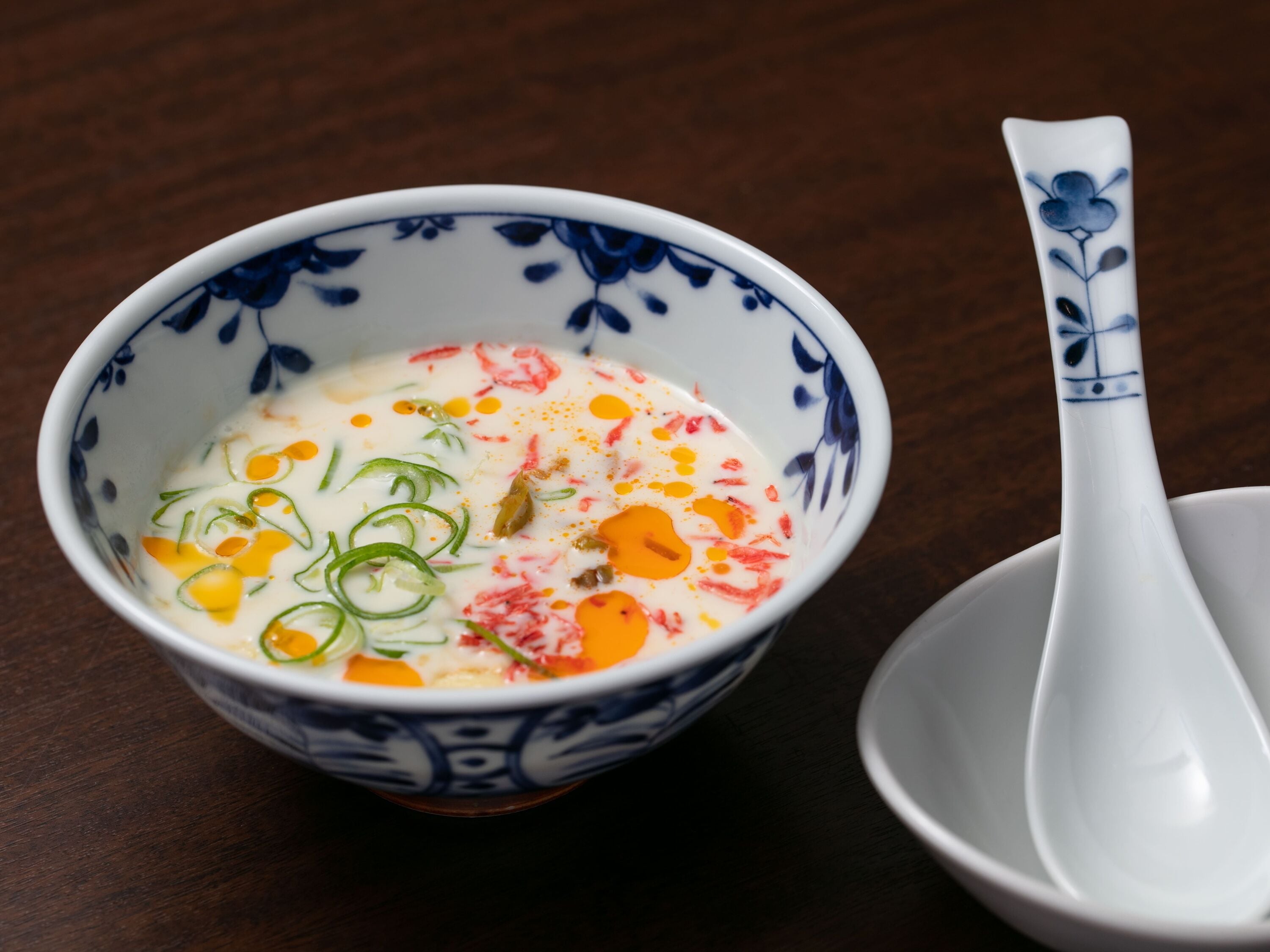 A bowl of soup with various ingredients, a spoon, and a decorative bowl on a wooden table.